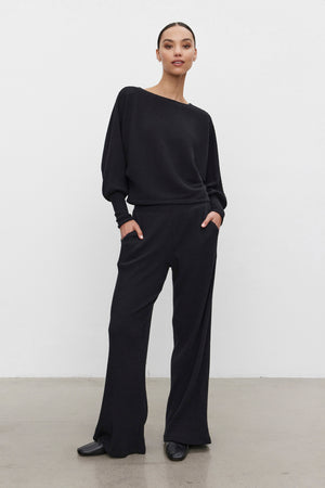 A person stands against a plain background wearing the COLLEEN RIBBED PANT and a matching black long-sleeve top from Velvet by Graham & Spencer, both made from ultra-soft fabric, with hands in pockets.