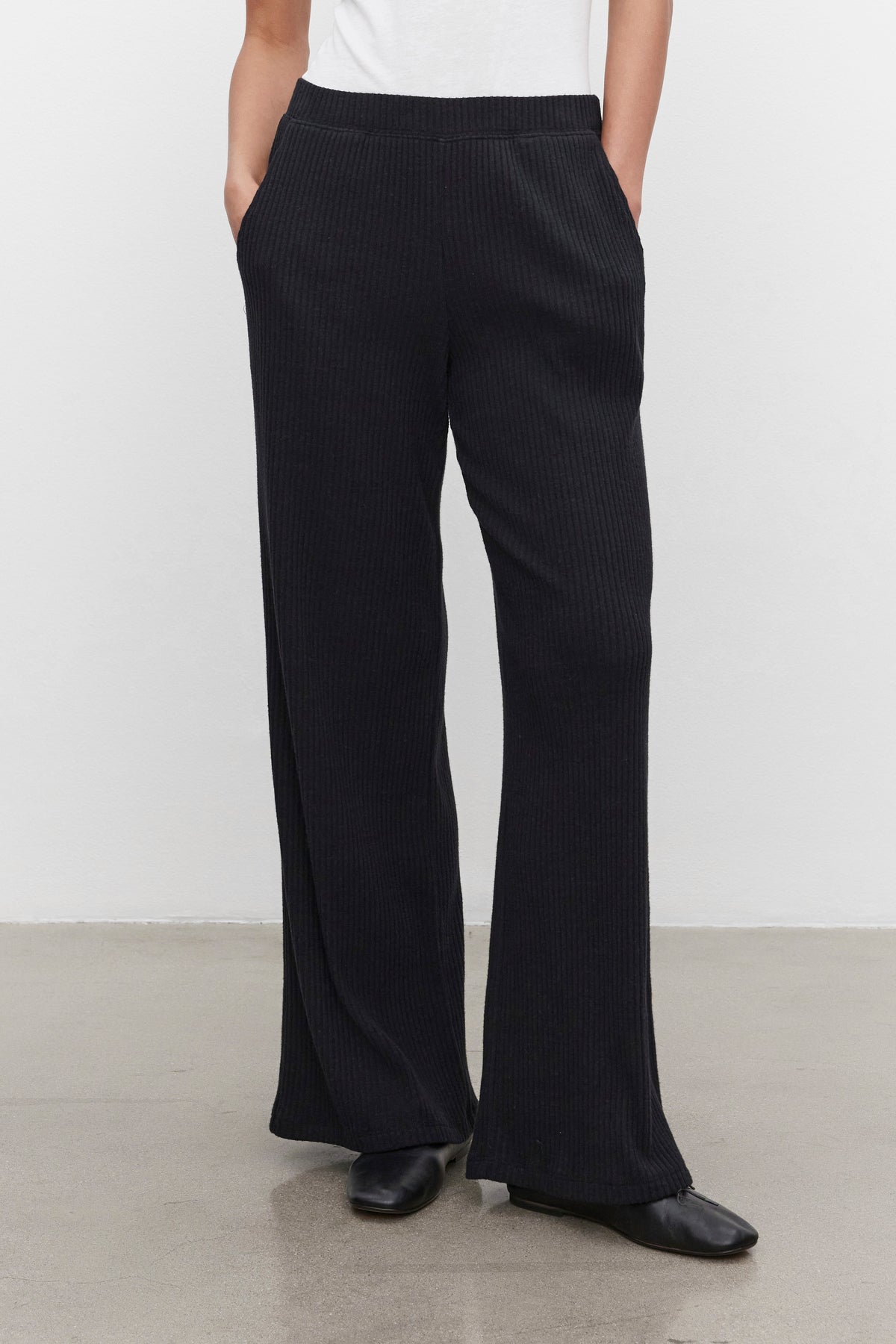   A person is wearing the COLLEEN RIBBED PANT by Velvet by Graham & Spencer in black, featuring a wide-leg silhouette and crafted from ultra-soft fabric. They pair it with a white top and stand against a plain white and gray background with their hands in their pockets. 