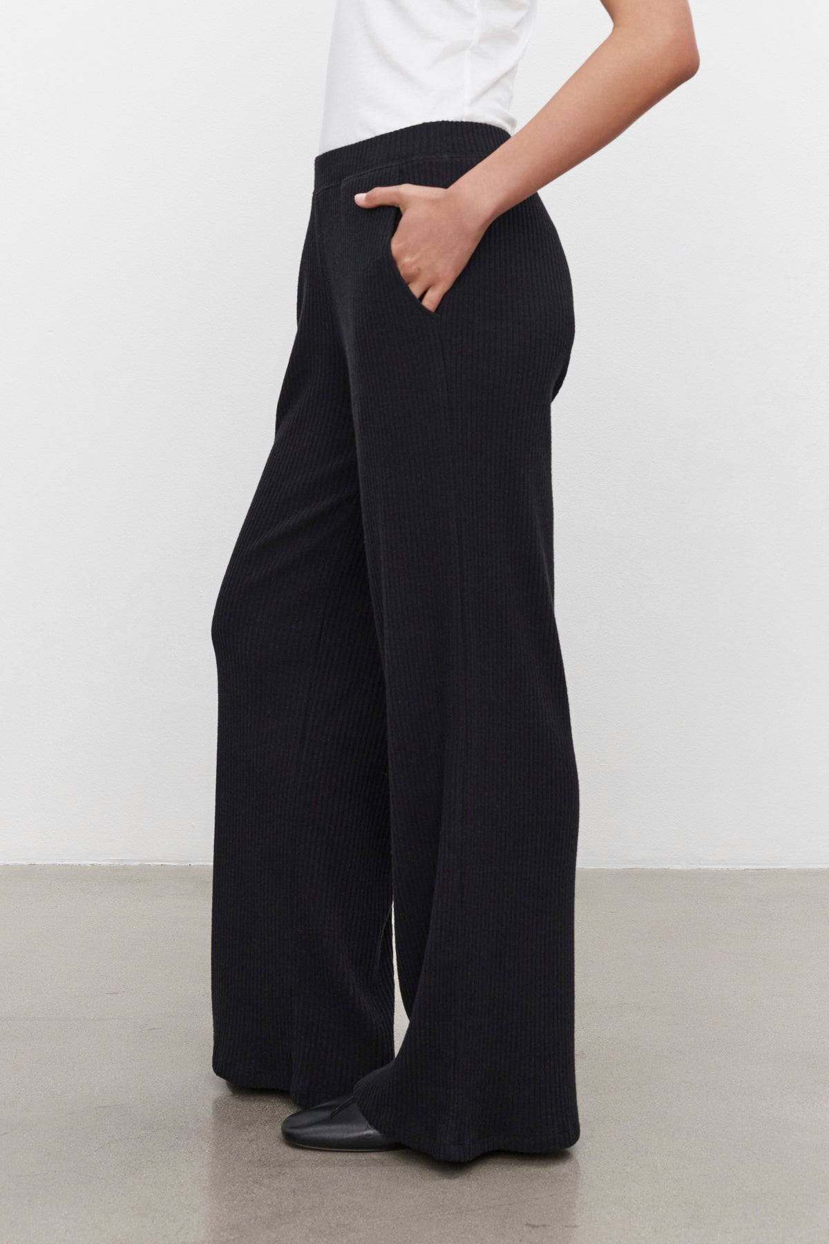   Person wearing Velvet by Graham & Spencer's COLLEEN RIBBED PANT in black and a white top, with one hand in their pocket. The photo is taken from a side angle and shows from the shoulder down. The plain white background highlights the sleek, everyday wear ensemble. 