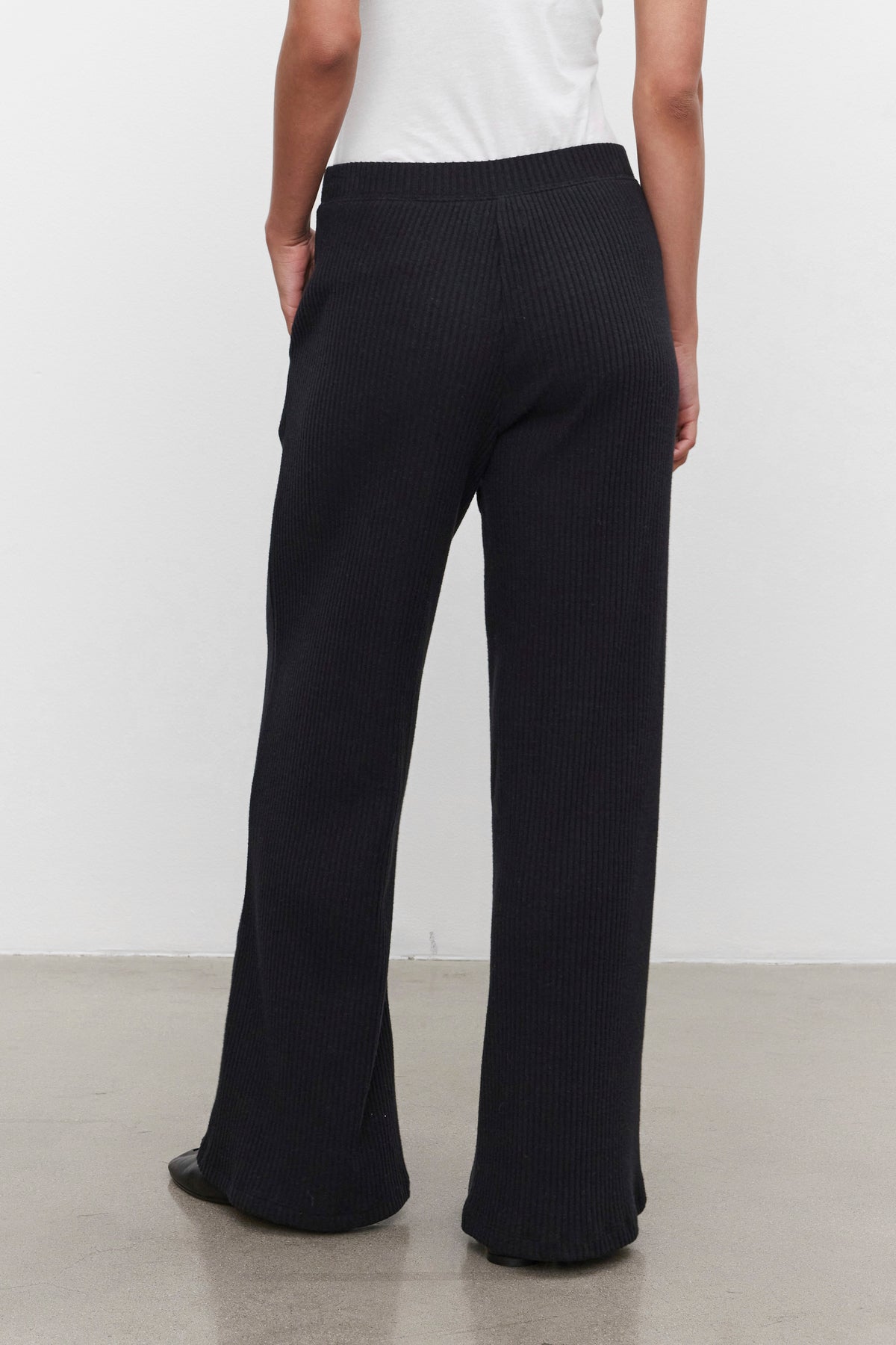   A person stands against a plain wall, wearing the ultra-soft COLLEEN RIBBED PANT in black from Velvet by Graham & Spencer, paired with a white top. The image, taken from behind, highlights the everyday wear appeal of these wide-leg pants. 