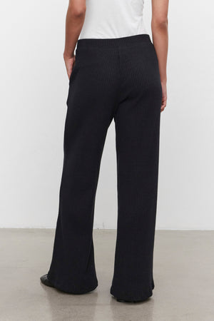 A person stands against a plain wall, wearing the ultra-soft COLLEEN RIBBED PANT in black from Velvet by Graham & Spencer, paired with a white top. The image, taken from behind, highlights the everyday wear appeal of these wide-leg pants.