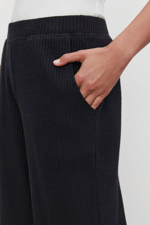 A person wearing the ultra-soft COLLEEN RIBBED PANT in black by Velvet by Graham & Spencer, with their right hand in the pocket, showcasing a stylish yet comfortable piece of everyday wear.