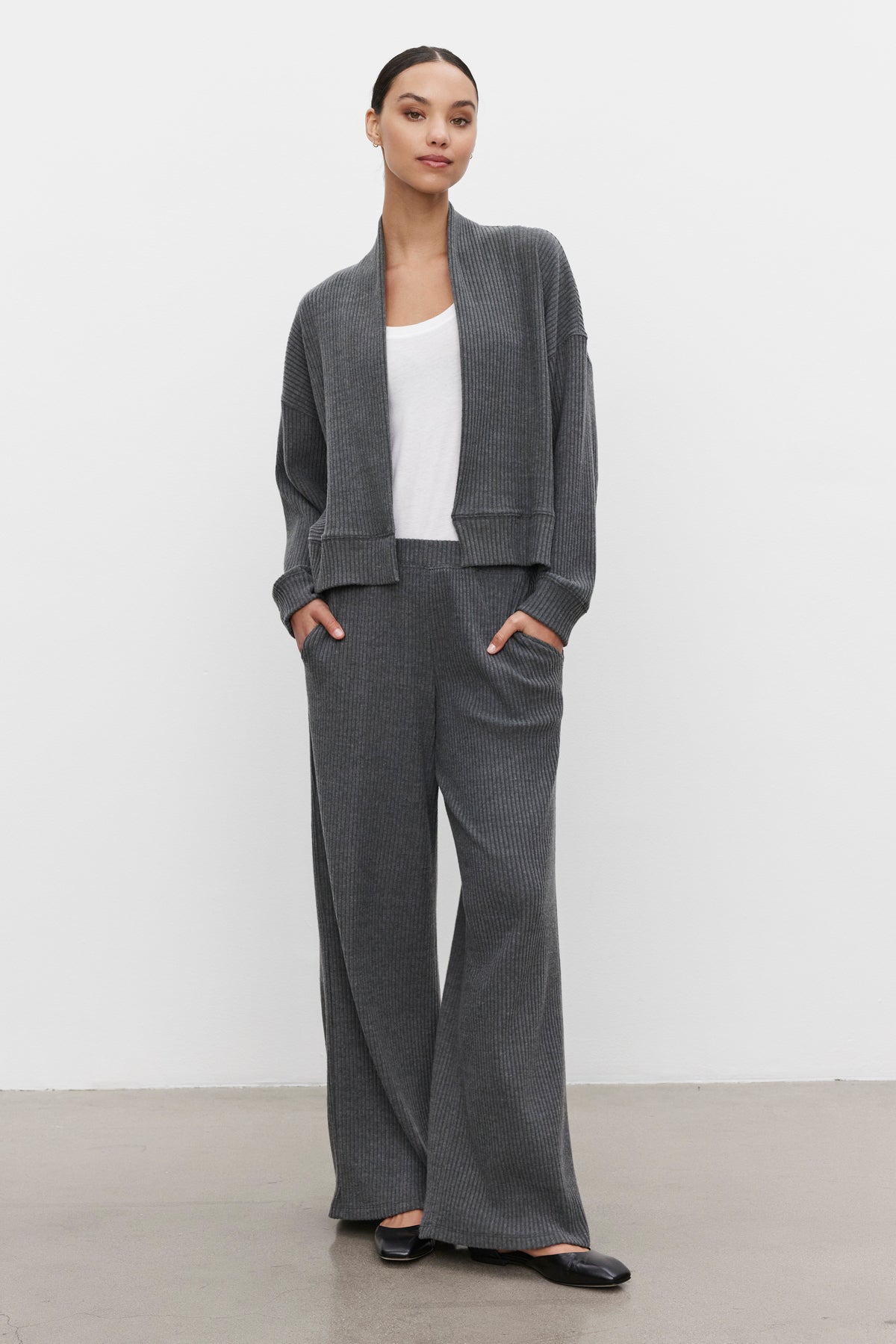   A person is standing in ultra-soft fabric, wearing a gray knit cardigan and matching COLLEEN RIBBED PANT by Velvet by Graham & Spencer with a white top, hands in pockets. The background is plain and white—perfect for everyday wear. 