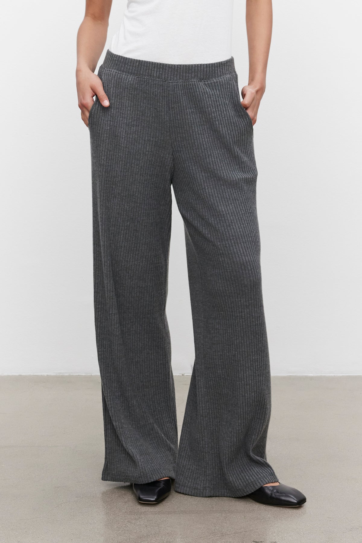   A person is wearing the ultra-soft COLLEEN RIBBED PANT by Velvet by Graham & Spencer in a stylish grey wide-leg design, paired with a white top, hands comfortably in pockets, and black shoes—perfect for everyday wear. 