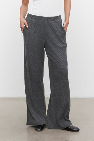 A person is wearing the ultra-soft COLLEEN RIBBED PANT by Velvet by Graham & Spencer in a stylish grey wide-leg design, paired with a white top, hands comfortably in pockets, and black shoes—perfect for everyday wear.