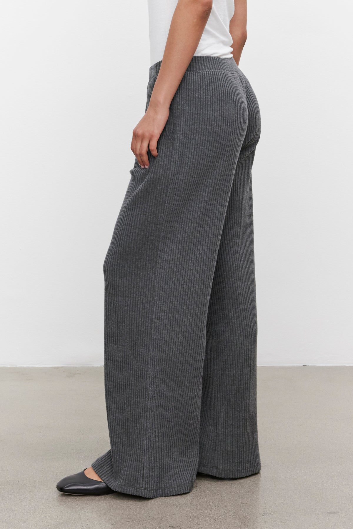   A person wearing a white top and the COLLEEN RIBBED PANT from Velvet by Graham & Spencer, made of ultra-soft fabric, is standing on a light gray floor, facing slightly away from the camera. 