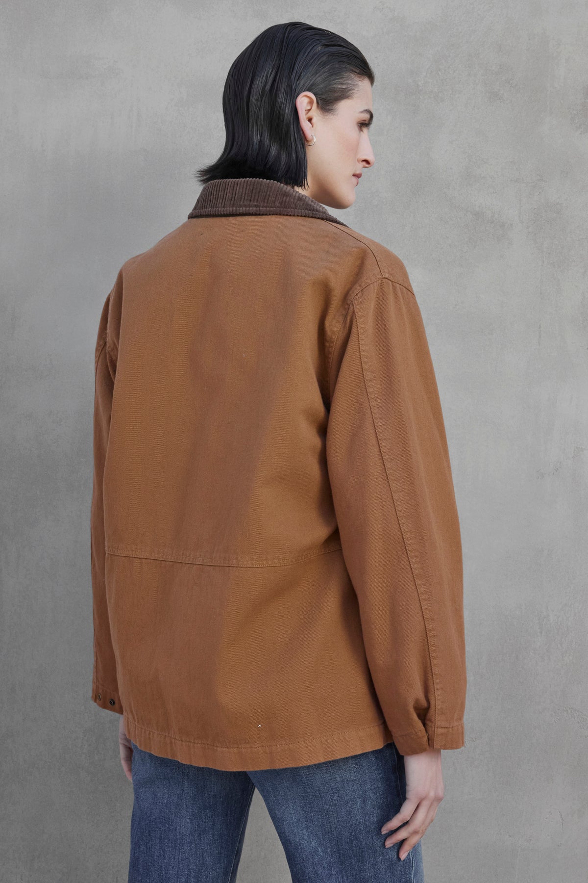   A person with short dark hair is seen facing away from the camera, wearing the OAKLEY JACKET by Velvet by Jenny Graham, which features corduroy trims, and pairing it with blue jeans while standing against a gray textured wall. 