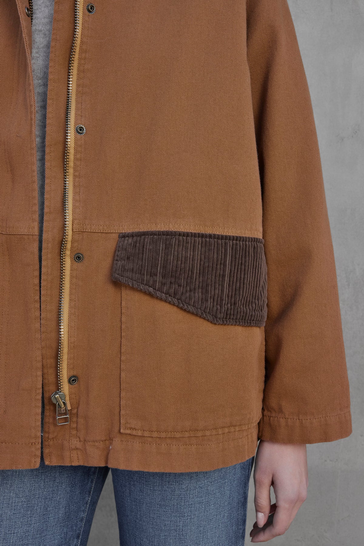   Close-up of a person's torso wearing the OAKLEY JACKET by Velvet by Jenny Graham. The brown jacket, made from durable cotton twill fabric, features a secure zipper front and contrasting corduroy trims on the pocket. It pairs perfectly with blue jeans. 