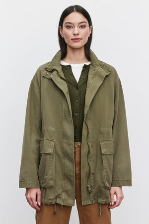 A person wearing the ODETTE JACKET by Velvet by Graham & Spencer in a durable cotton twill olive green with a zipper front over a green cardigan and light brown pants stands against a plain white background.