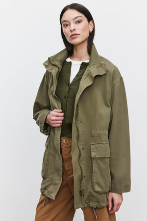 A woman with long dark hair wears the ODETTE JACKET from Velvet by Graham & Spencer, made of durable cotton twill in olive green. The jacket features a classic collar and zipper front, layered over a green sweater and white top. She pairs it with brown pants and stands against a plain white background, looking at the camera.