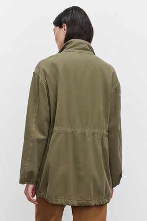 A person with long dark hair is turned away from the camera, showcasing the ODETTE JACKET by Velvet by Graham & Spencer. The olive green jacket, crafted from durable cotton twill, features a high collar and an adjustable waist. The individual pairs it with brown pants against a plain white background.