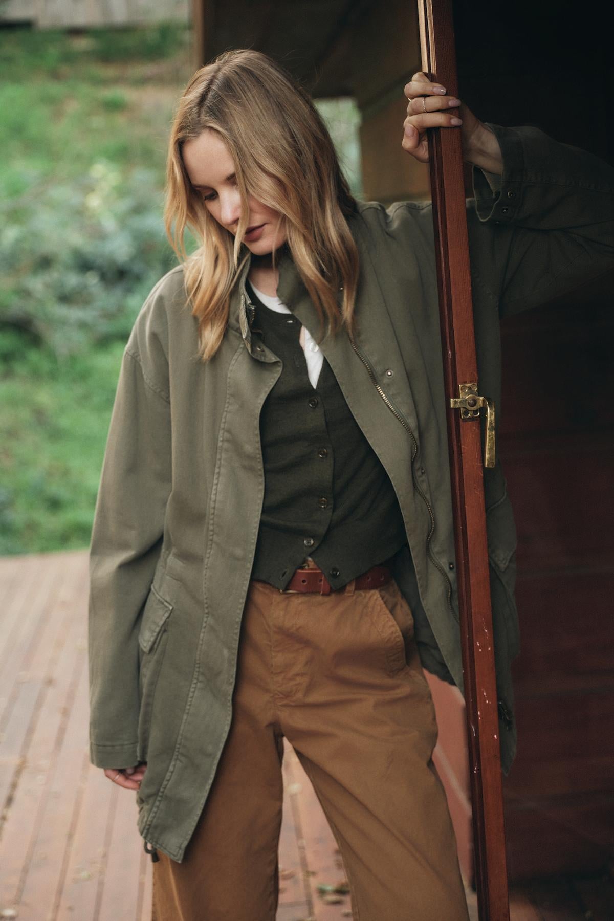   A woman in casual clothing, featuring an oversized jacket and the luxurious VIOLETTE CARDIGAN from Velvet by Graham & Spencer, stands in a doorway with her hand on the doorframe, looking downward outside a wooden structure. 