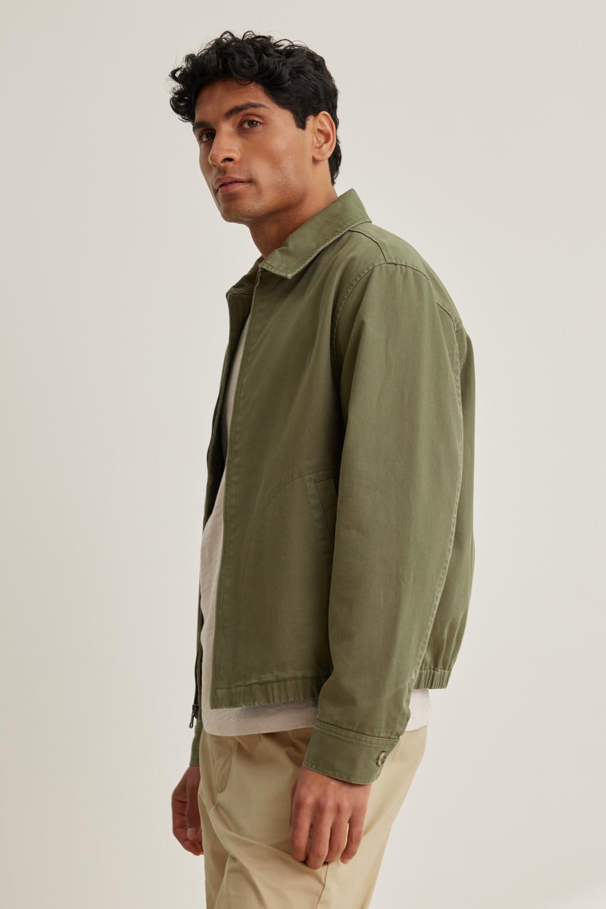 A person stands sideways in an olive COLLINS JACKET by Velvet by Graham & Spencer, paired with a white shirt and beige pants against a neutral background.-38643251413185