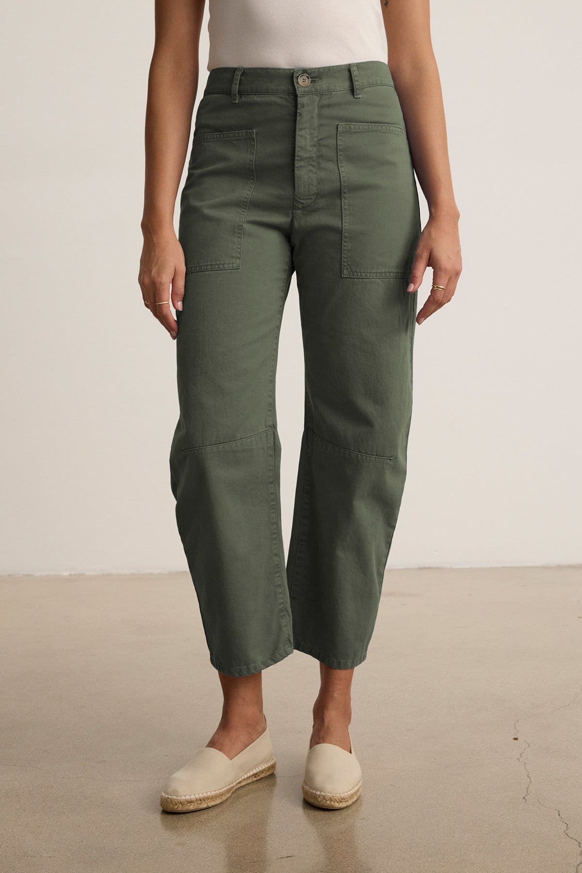 A person stands on a beige floor by a white wall, wearing Velvet by Graham & Spencer's olive-green BRYLIE SANDED TWILL UTILITY PANT made from cotton twill. Their look is completed with beige espadrilles for an effortless style.-38807322656961