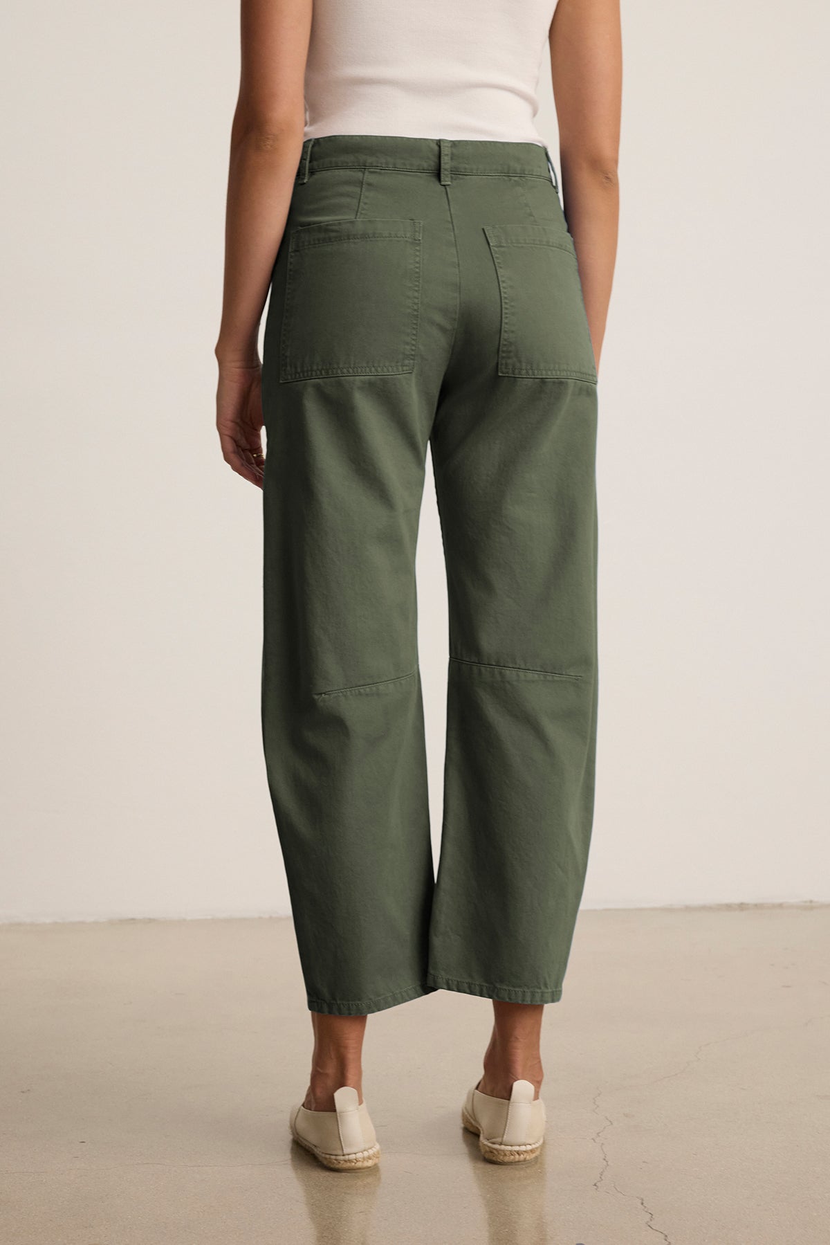   A person wearing a white top and the BRYLIE SANDED TWILL UTILITY PANT by Velvet by Graham & Spencer, featuring green wide legs and patch pockets, stands on a plain floor, facing away. 