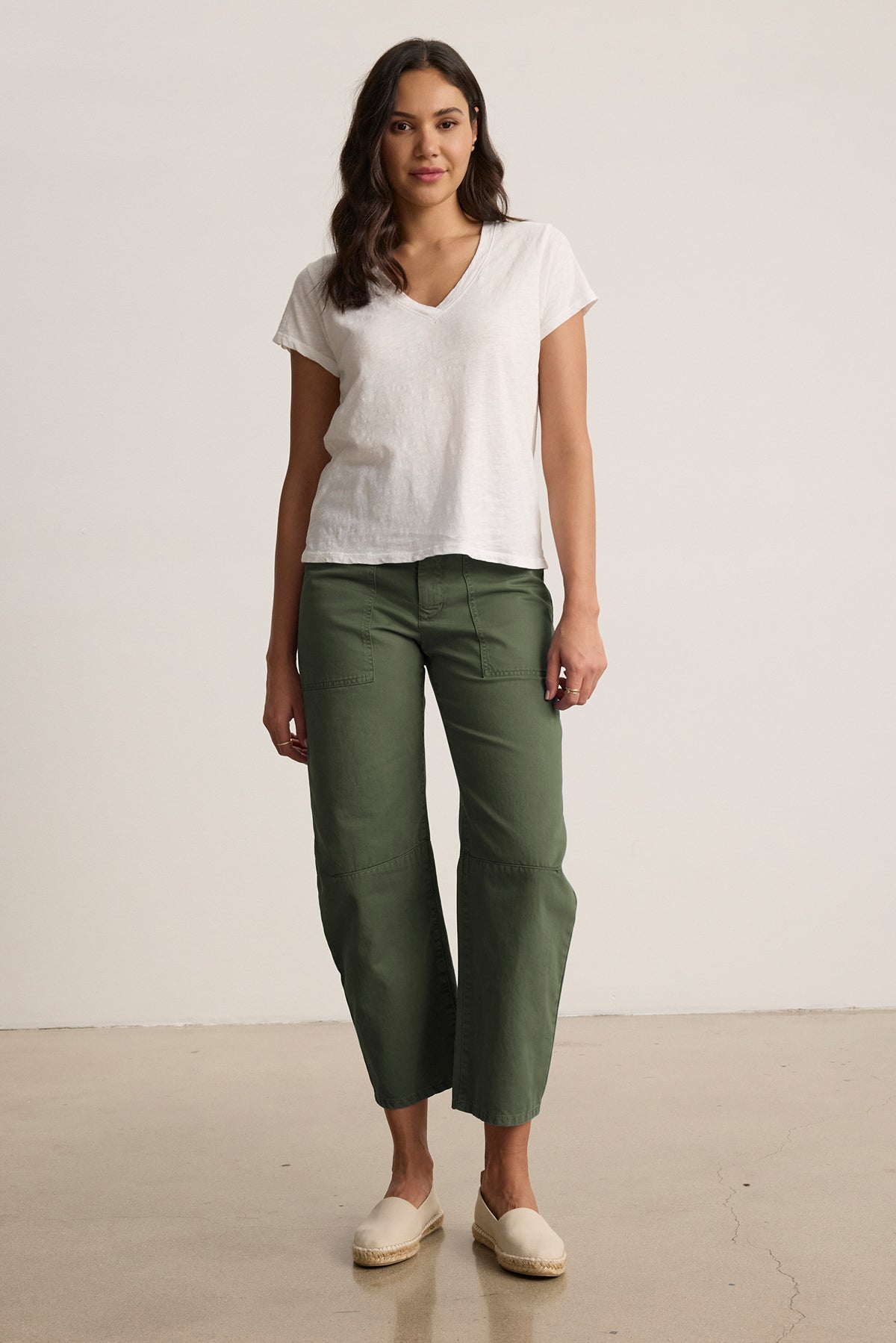   A person stands against a plain background wearing a white V-neck t-shirt, Velvet by Graham & Spencer's BRYLIE SANDED TWILL UTILITY PANT with patch pockets, and beige slip-on shoes. 