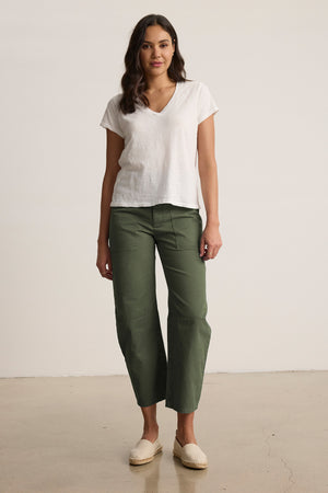 A person stands against a plain background wearing a white V-neck t-shirt, Velvet by Graham & Spencer's BRYLIE SANDED TWILL UTILITY PANT with patch pockets, and beige slip-on shoes.