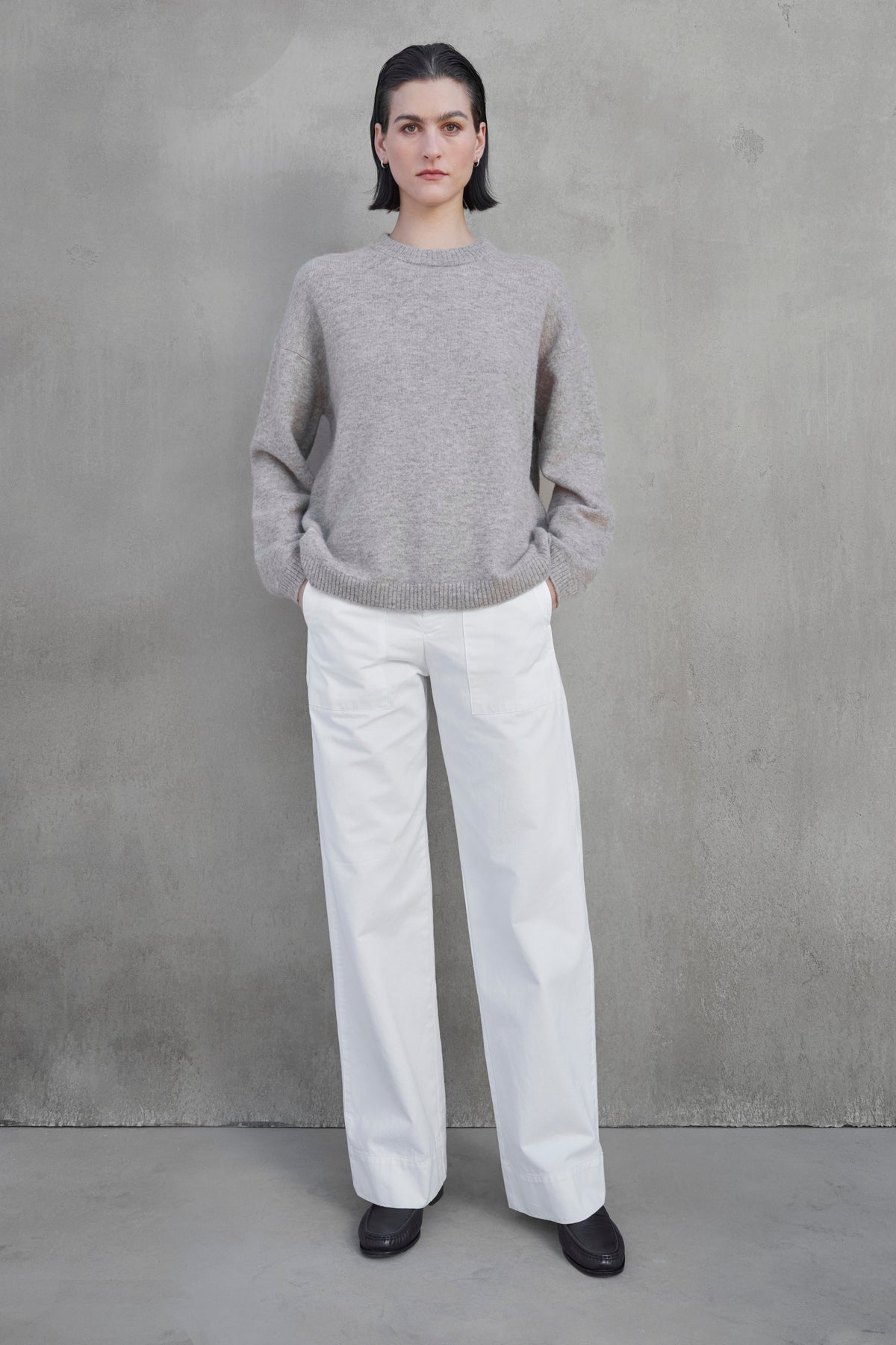 A person with short dark hair stands against a gray wall, wearing the LAKEWOOD ALPACA SWEATER by Velvet by Jenny Graham, white wide-leg pants, and black shoes. The person's hands are tucked into their pockets.-37659909259457