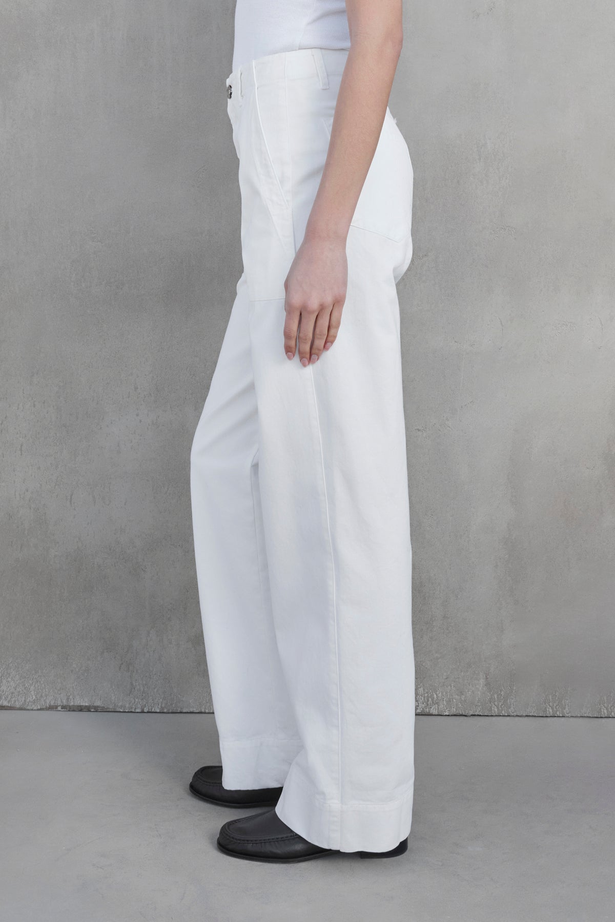 Side view of a person wearing the VENTURA PANT by Velvet by Jenny Graham, styled with a white top and black shoes, standing against a gray textured wall.-37659921875137