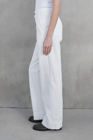 Side view of a person wearing the VENTURA PANT by Velvet by Jenny Graham, styled with a white top and black shoes, standing against a gray textured wall.