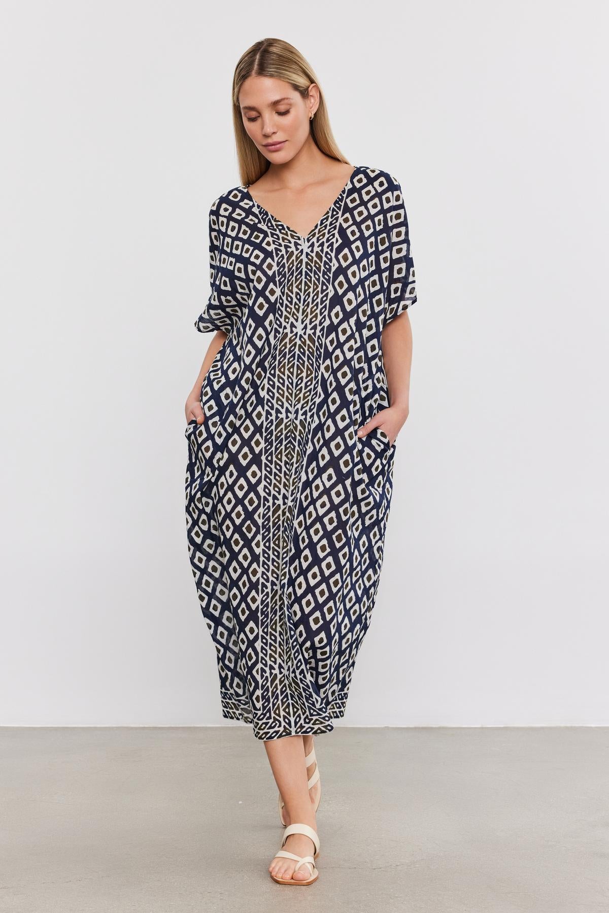   A woman stands against a plain white background, wearing the ODESSA KAFTAN DRESS from Velvet by Graham & Spencer. This blue and white geometric-patterned dress features a v-neckline and short sleeves. Made from lightweight cotton, this stylish kaftan allows her to place her hands comfortably in the dress pockets and pairs perfectly with her sandals—an ideal addition to any vacation wardrobe. 