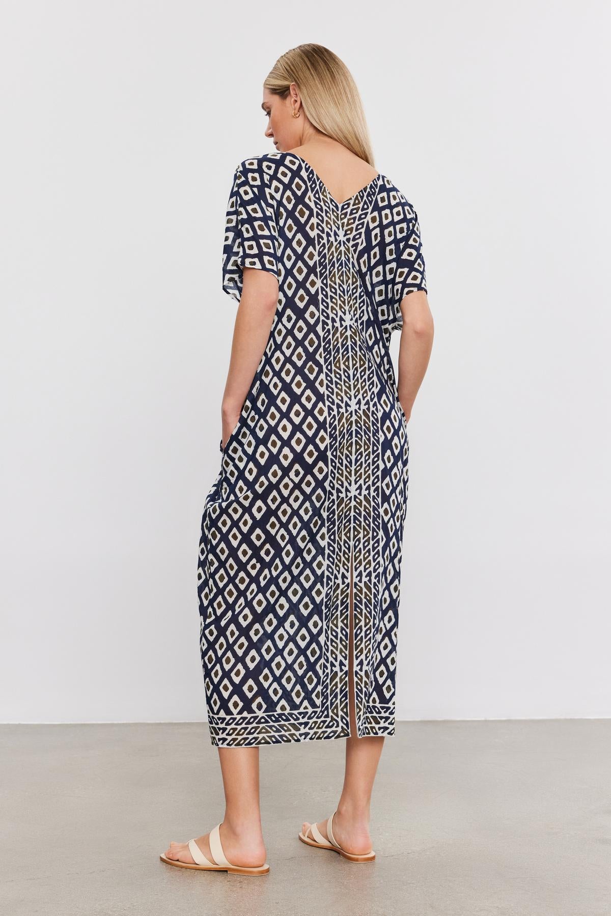   A woman in a Velvet by Graham & Spencer ODESSA KAFTAN DRESS featuring geometric designs, standing with her back to the camera. She is wearing sandals, showcasing a perfect addition to any vacation wardrobe in lightweight cotton. 