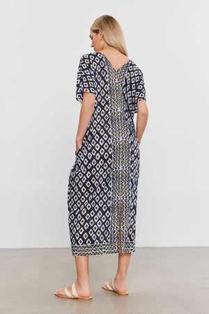 A woman in a Velvet by Graham & Spencer ODESSA KAFTAN DRESS featuring geometric designs, standing with her back to the camera. She is wearing sandals, showcasing a perfect addition to any vacation wardrobe in lightweight cotton.