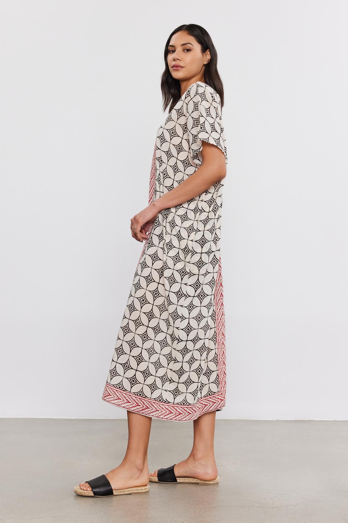   A woman stands in profile, wearing a lightweight cotton Odessa kaftan dress by Velvet by Graham & Spencer with short sleeves and sandals, on a plain white background. 