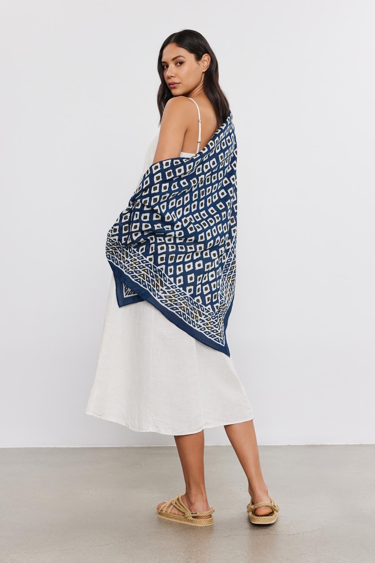   A woman in a white dress and espadrilles, draped in a Velvet by Graham & Spencer SARONG WRAP, stands looking over her shoulder against a plain background. 