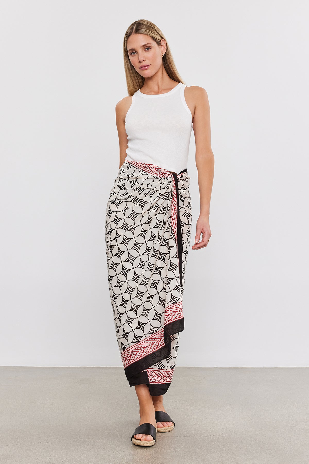   Woman wearing a white sleeveless top and a SARONG WRAP by Velvet by Graham & Spencer, standing on a beige floor against a plain white background. 