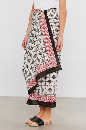 Person wearing a Velvet by Graham & Spencer SARONG WRAP with black and red borders, reminiscent of a sarong, stands on a gray floor, paired with a white top and black slide sandals.