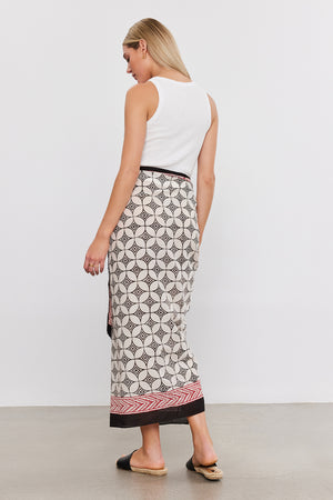 A person with long blonde hair, wearing a white sleeveless top and a Velvet by Graham & Spencer SARONG WRAP featuring vibrant prints, stands with their back towards the camera in a minimalist indoor setting.