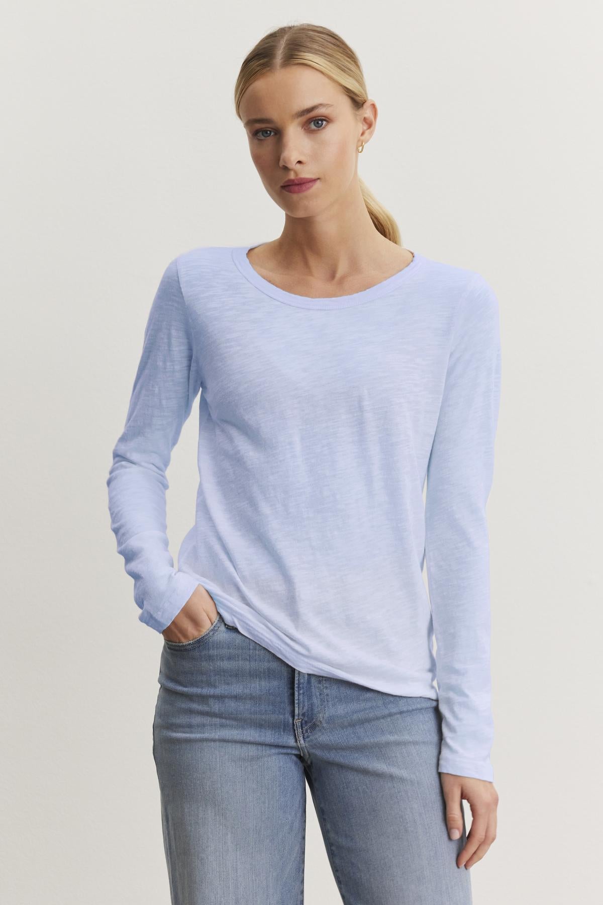   A person in a Velvet by Graham & Spencer LIZZIE TEE, featuring a light blue long-sleeve design with a crew neckline made from textured cotton slub, stands against a plain backdrop with one hand in their pocket, stylishly paired with jeans. 