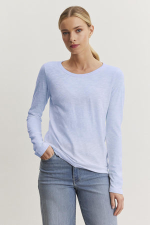 A person in a Velvet by Graham & Spencer LIZZIE TEE, featuring a light blue long-sleeve design with a crew neckline made from textured cotton slub, stands against a plain backdrop with one hand in their pocket, stylishly paired with jeans.