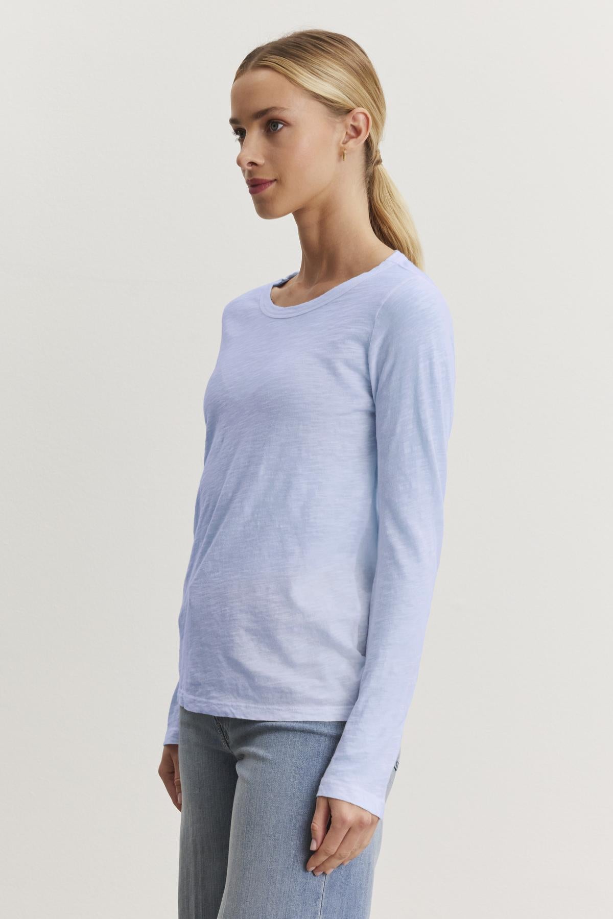   Wearing Velvet by Graham & Spencer's LIZZIE TEE in light blue, featuring textured cotton slub with a crew neckline and long sleeves, a person pairs it with jeans and gazes thoughtfully to the side against a plain background. 