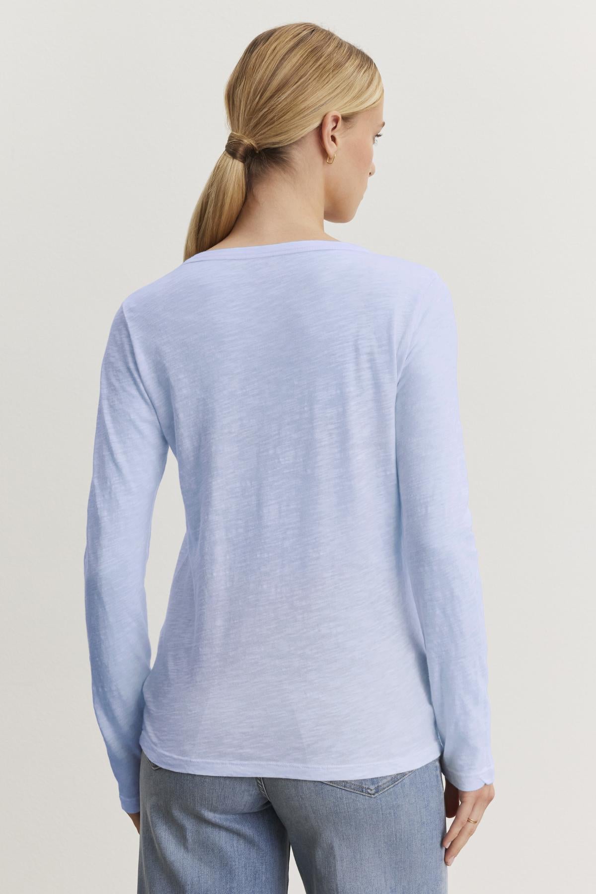   Person with long hair in a ponytail wearing the LIZZIE TEE by Velvet by Graham & Spencer, a light blue, textured cotton slub shirt with a crew neckline and long sleeves, paired with jeans, facing away. 