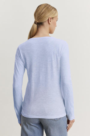 Person with long hair in a ponytail wearing the LIZZIE TEE by Velvet by Graham & Spencer, a light blue, textured cotton slub shirt with a crew neckline and long sleeves, paired with jeans, facing away.