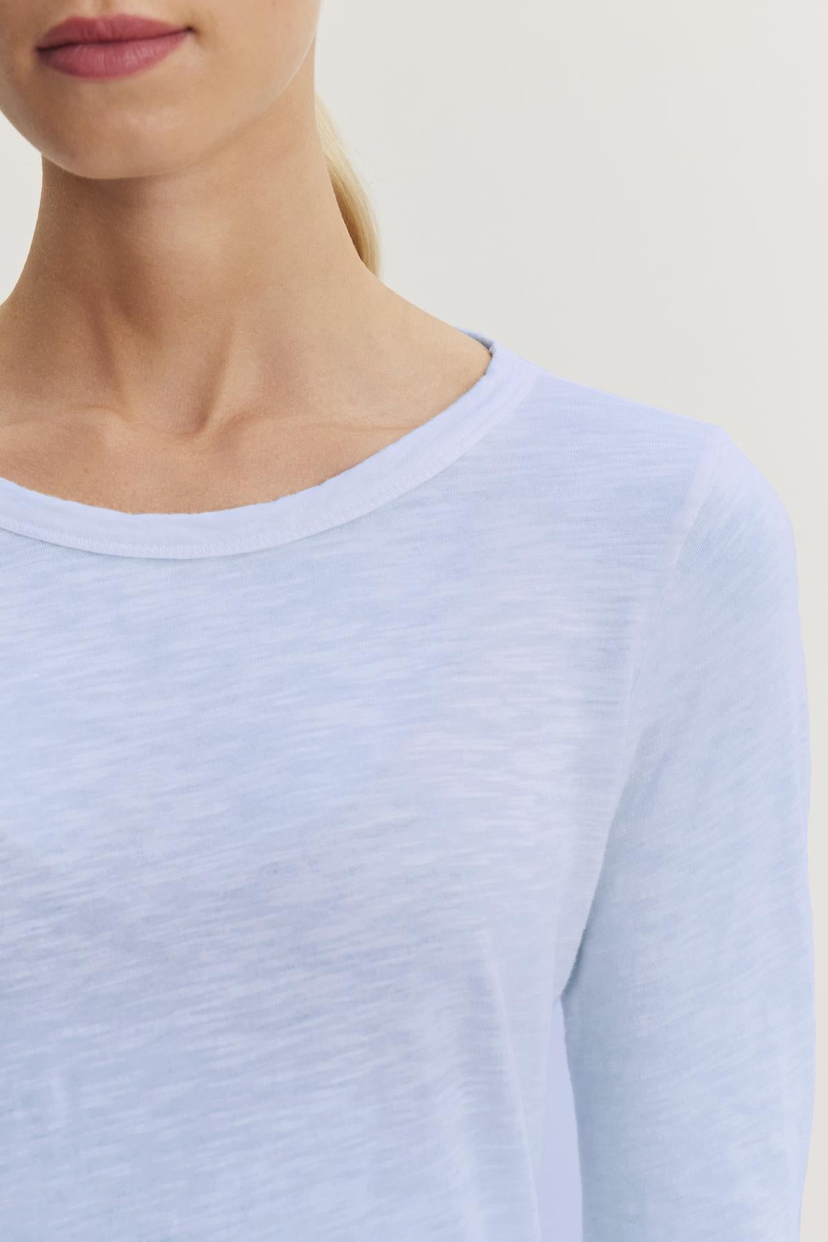   Someone is wearing the Velvet by Graham & Spencer LIZZIE TEE, a light blue long-sleeve shirt with a classic crew neckline. The textured cotton slub provides subtle depth against a plain background, visible from neck to shoulder. 