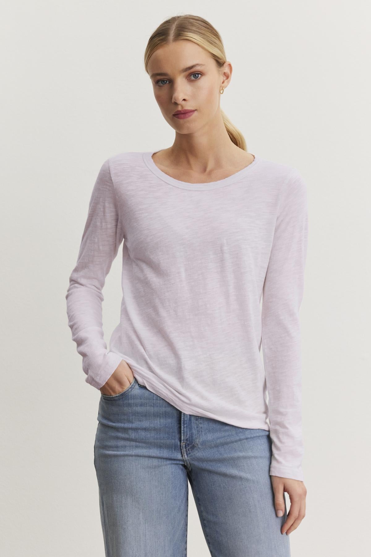   A person in the LIZZIE TEE by Velvet by Graham & Spencer, a light pink long-sleeve shirt with a classic crew neckline, and blue jeans stands against a plain white background. 