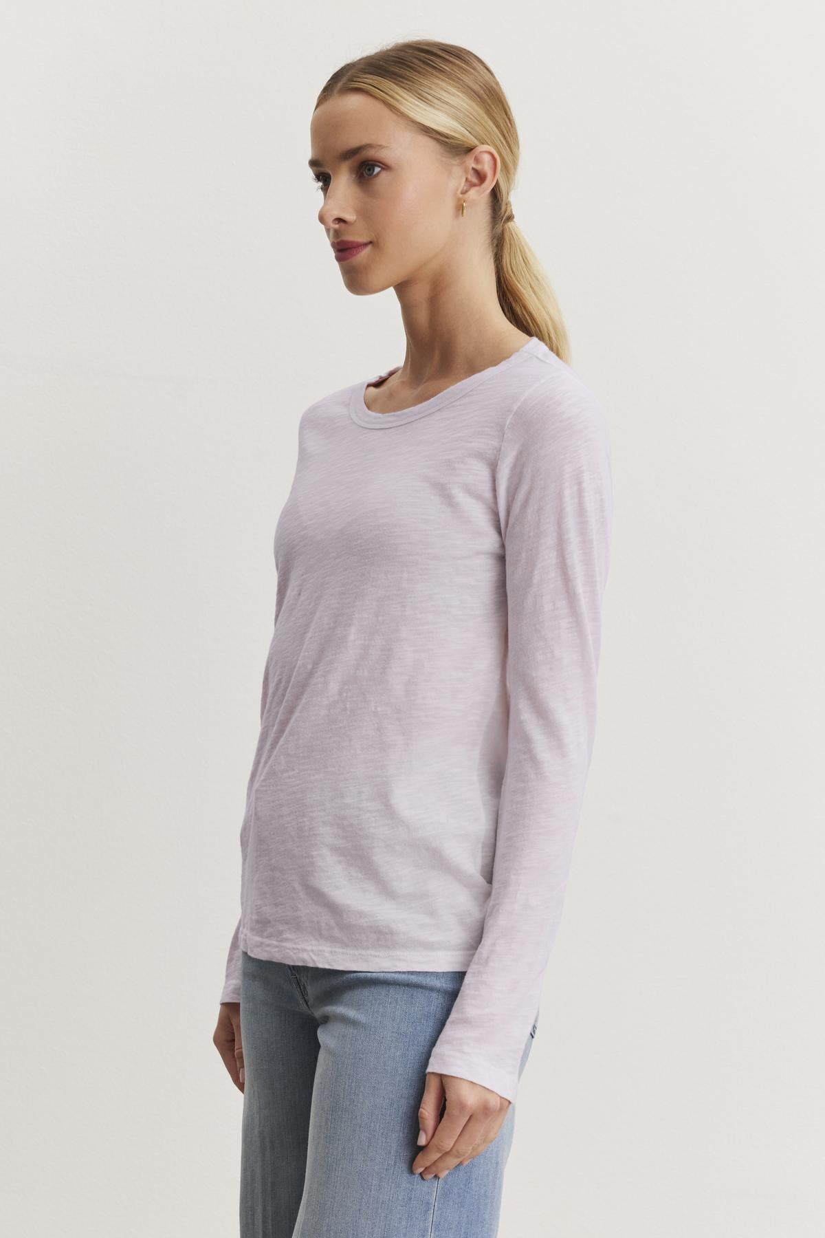   A person wearing the Velvet by Graham & Spencer LIZZIE TEE in light pink, featuring long sleeves and a classic crew neckline, paired with jeans, stands facing left against a plain white background. 