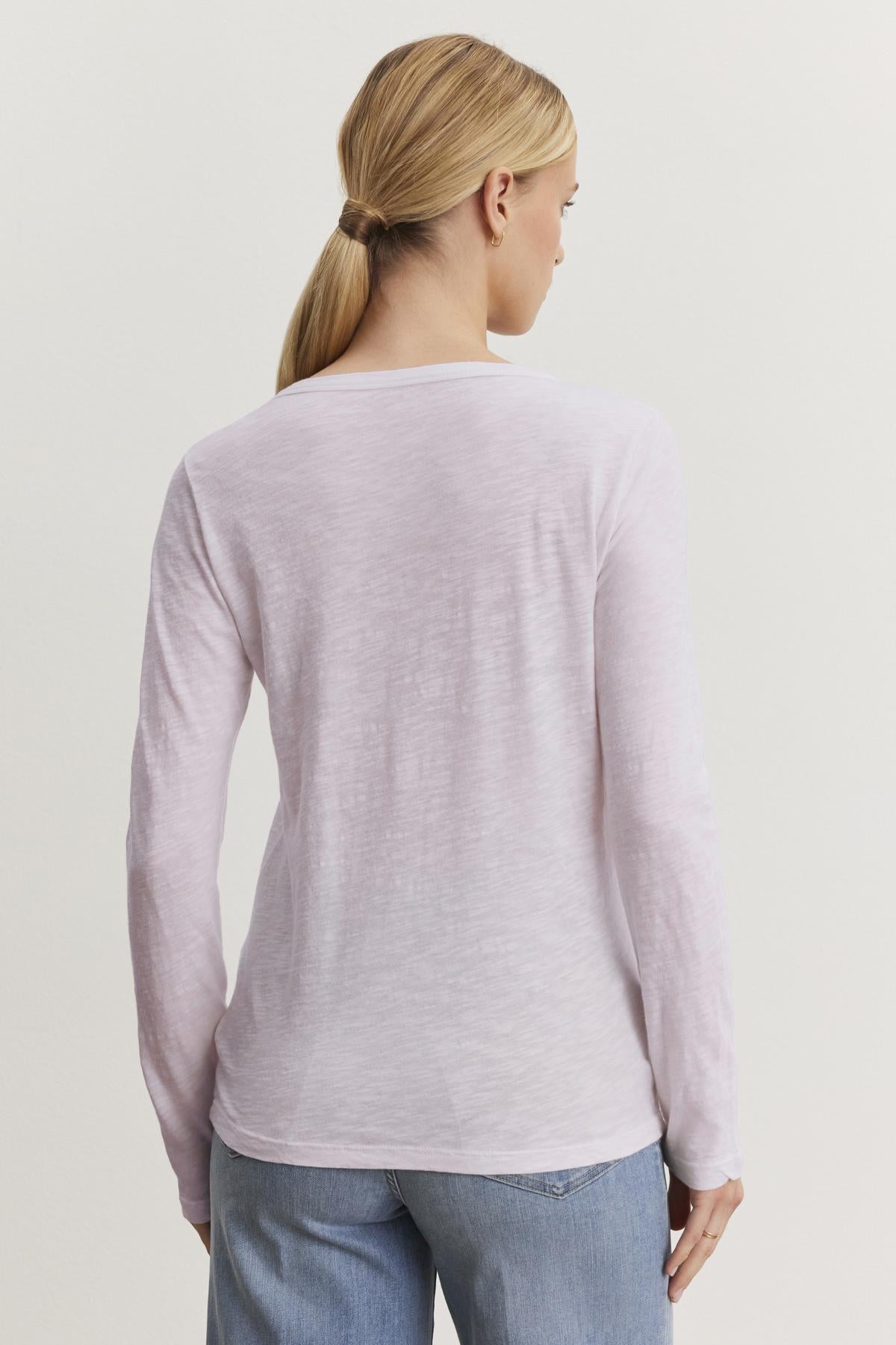   A person with long blonde hair in a ponytail, wearing the LIZZIE TEE—a light pink long-sleeve shirt made from textured cotton slub by Velvet by Graham & Spencer—and blue jeans, faces away against a plain background. 