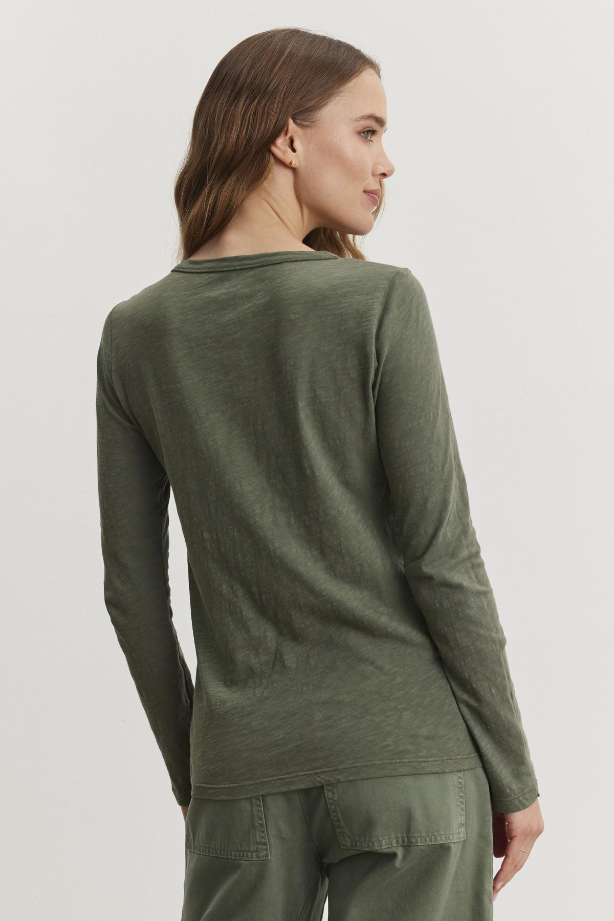   A person with long hair is wearing Velvet by Graham & Spencer's LIZZIE TEE, an olive green cotton slub top, along with pants. The outfit features a subtle crew neckline as they face away. 