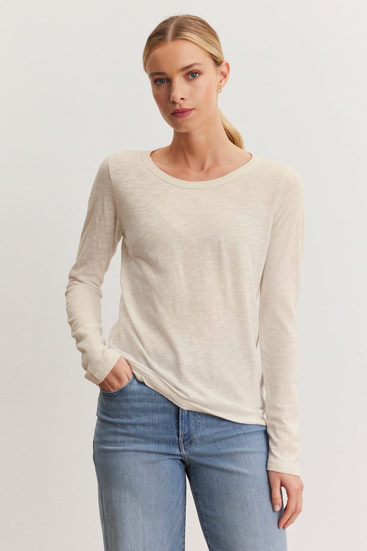 A person poses against a neutral background in blue jeans and the Velvet by Graham & Spencer LIZZIE TEE, a cream long-sleeve shirt with a classic crew neckline, with one hand in their pocket.-38662713802945