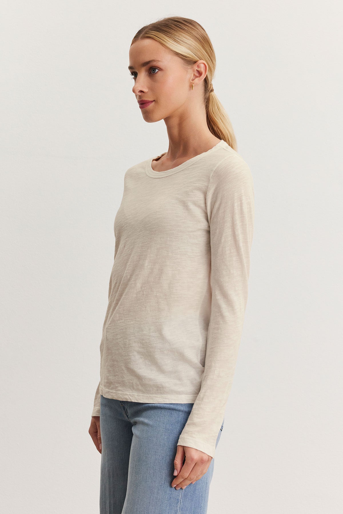   A person with light hair tied back is looking forward, wearing the LIZZIE TEE by Velvet by Graham & Spencer—a cream long-sleeve shirt with a crew neckline—and light blue jeans, standing against a plain light background. The textured cotton slub adds subtle sophistication to the casual outfit. 