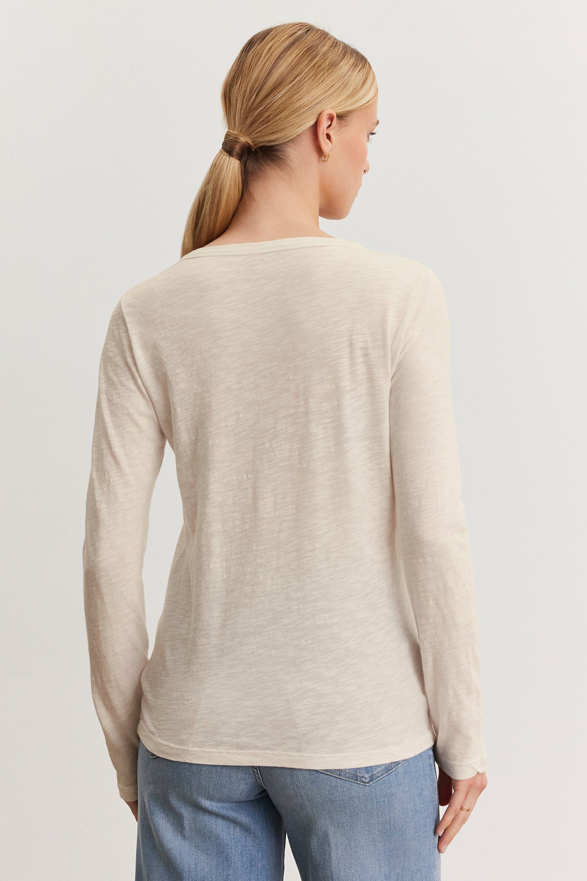 A person with long hair in a ponytail wears the LIZZIE TEE by Velvet by Graham & Spencer, featuring a beige textured cotton slub long-sleeve design, paired with blue jeans, facing away from the camera against a plain background.-38662713835713