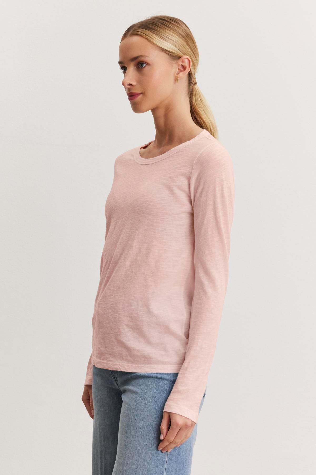 A person with a blond ponytail stands against a plain white background, wearing the LIZZIE TEE, a light pink cotton slub long sleeve top by Velvet by Graham & Spencer with a crew neckline, paired with blue jeans.-38301515251905