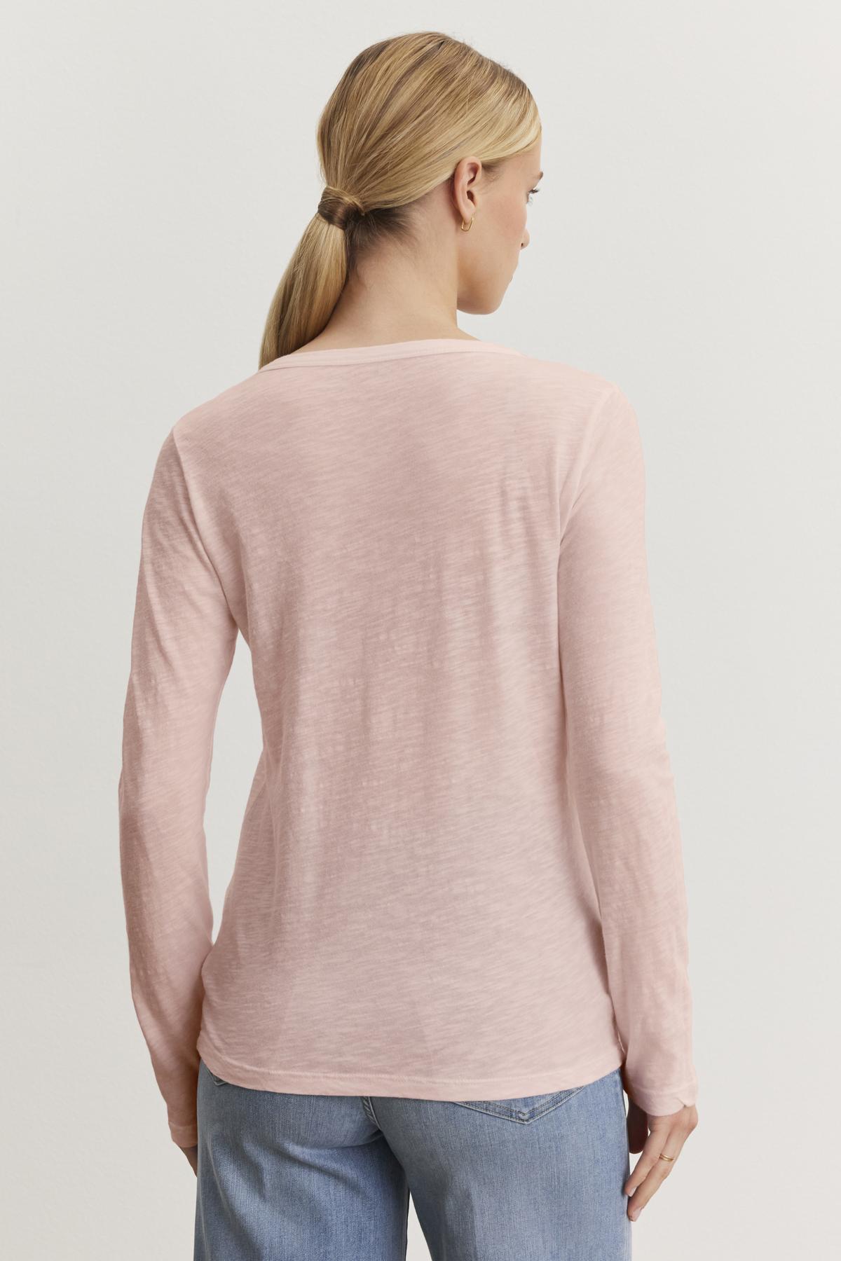 A person with long hair tied back is wearing the LIZZIE TEE, a light pink cotton slub long-sleeve top from Velvet by Graham & Spencer, paired with blue jeans, and is standing with their back to the camera against a plain background.-38301515317441