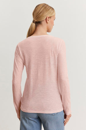 A person with long hair tied back is wearing the LIZZIE TEE, a light pink cotton slub long-sleeve top from Velvet by Graham & Spencer, paired with blue jeans, and is standing with their back to the camera against a plain background.