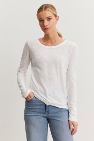 A person with blonde hair in a ponytail is wearing a white long-sleeve, classic crew neckline LIZZIE TEE by Velvet by Graham & Spencer and blue jeans, standing against a plain white background.