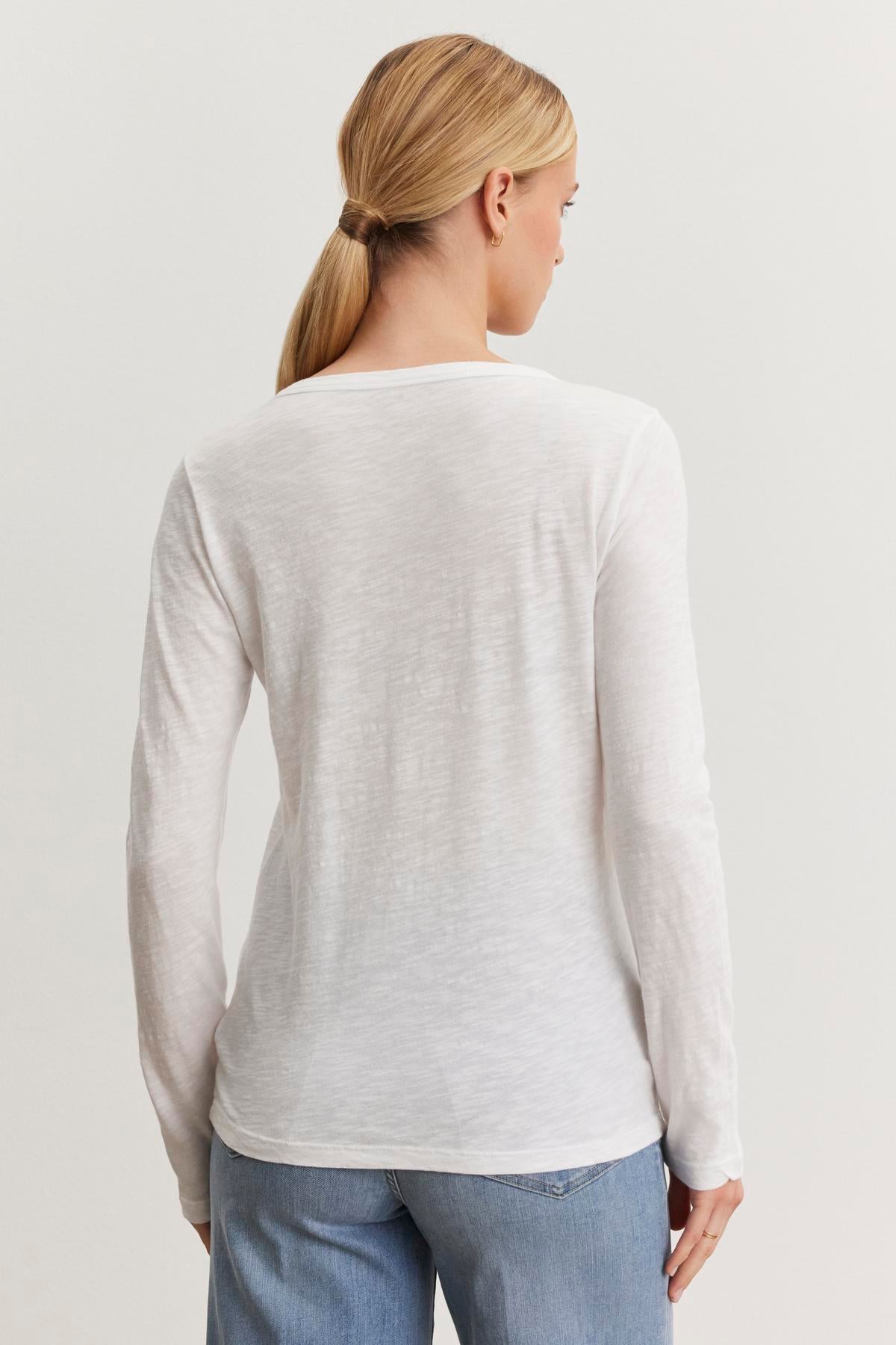 A person with blonde hair in a ponytail is shown from the back, wearing a long-sleeved white Velvet by Graham & Spencer LIZZIE TEE with a classic crew neckline and light blue jeans.-37366915203265